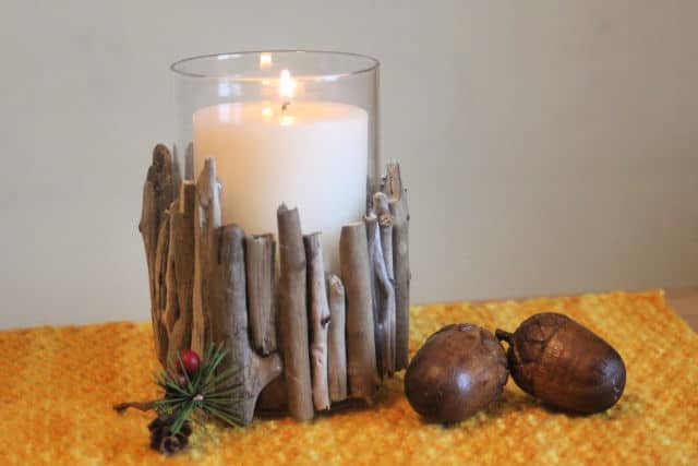 A glass candleholder decorated with pieces of driftwood sitting on an orange placemat surrounded by acorns.