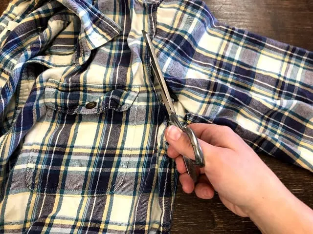 A hand with scissors cutting the sleeve off a flannel shirt.