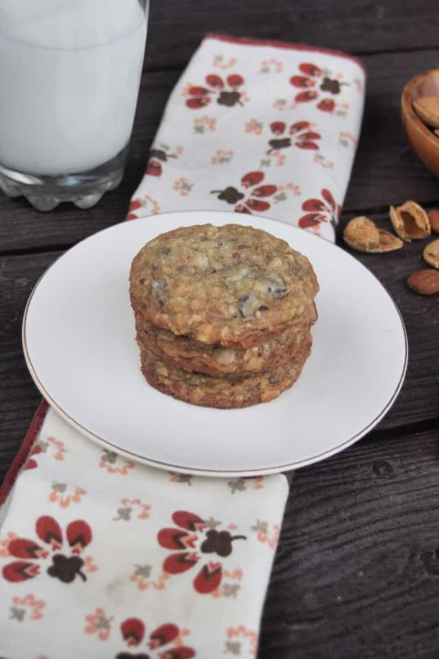 3 coconut chocolate chip cookies stacked on a white plate sitting on top of a napkin some almond shells and a glass of milk behind the plate.