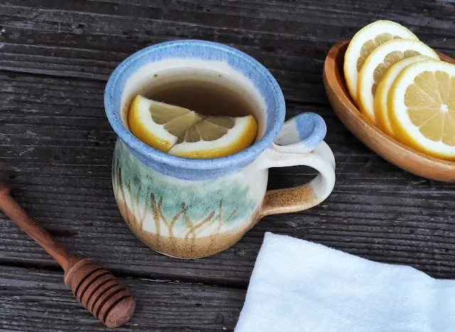 A cup of tea with lemon wedges surrounded by a honey dipper, white napkin, and wooden bowl of lemon slices. 