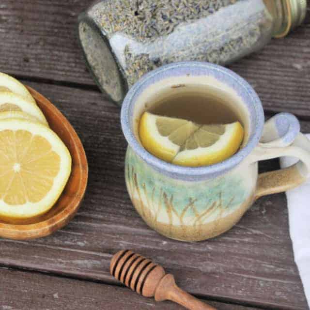 A cup of lavender hot toddy with lemons floating inside. Surrounded by a bowl of lemon slices, a honey dipper, a napkin, and a jar of dried lavender buds.
