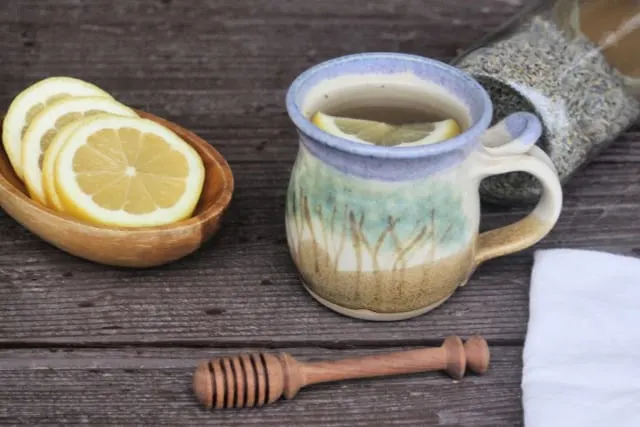 A cup of lavender tea surrounded by a jar of dried lavender, a bowl of lemon slices, a white napkin, and a honey dipper.