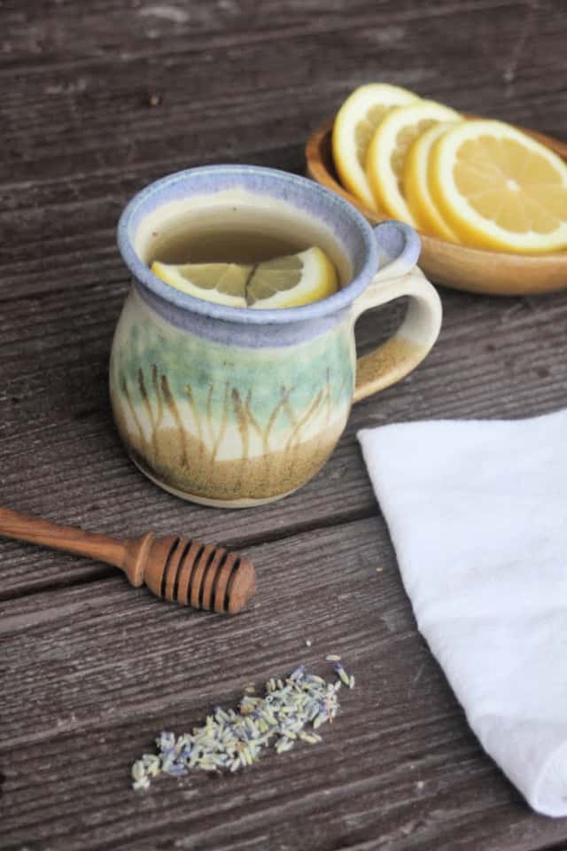 A mug of lavender hot toddy with lemon wedges, surrounded by dried lavender buds, a honey dipper, a white napkin, and a bowl of lemons on a wooden table.