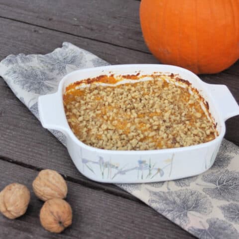 Pumpkin casserole in a baking dish sitting on top of a napkin surrounded by walnuts in shell and whole pumpkin.