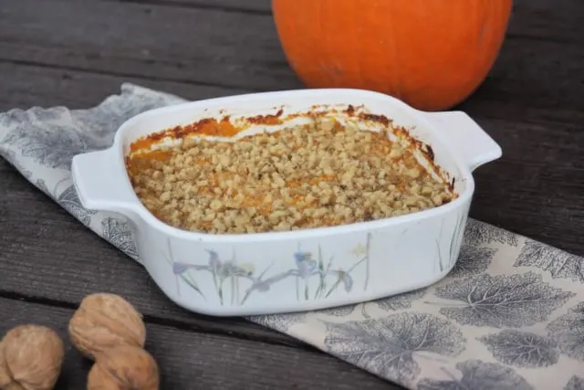 Pumpkin casserole in a baking dish sitting on top of a napkin surrounded by walnuts in shell and whole pumpkin.
