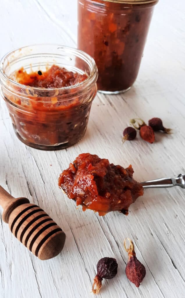 A spoon of dried rosehip jam with dried rosehips, honey dipper, and jars of jam on a table.