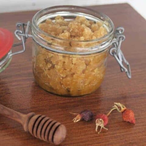 An open jar of rosehip sugar scrub on a table with honey dipper and dried rosehips.