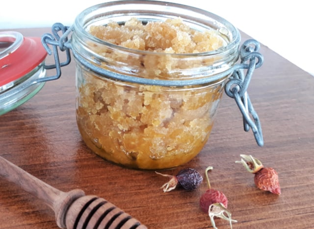 An open jar of rosehip sugar scrub on table seen from the side with a honey dipper and dried rosehips.