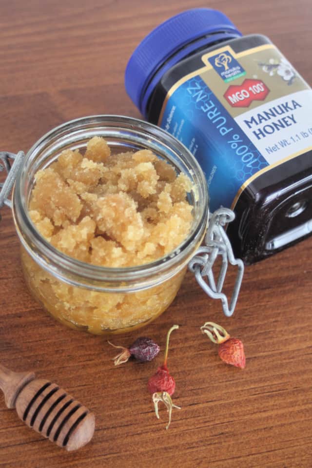 An open jar of rosehip sugar scrub on a table with a honey dipper, dried rosehips, and a jar of manuka honey.