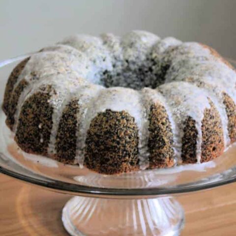 Glazed lemon poppy seed cake on clear glass cake stand.