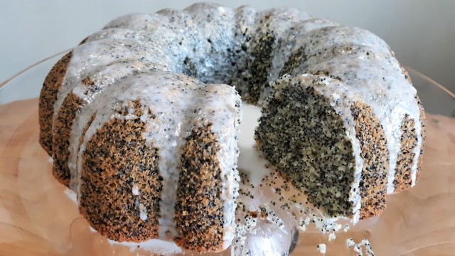 Glazed lemon poppy seed cake with a slice removed on glass cake plate. 