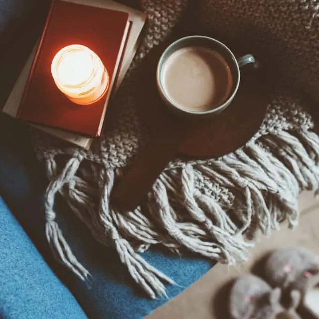 A coffee mug and books with lit candle sitting on blanket draped over a chair.