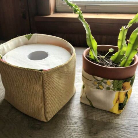 A fabric basket holding a roll of toilet paper sitting next to a potted plant in a floral fabric basket.