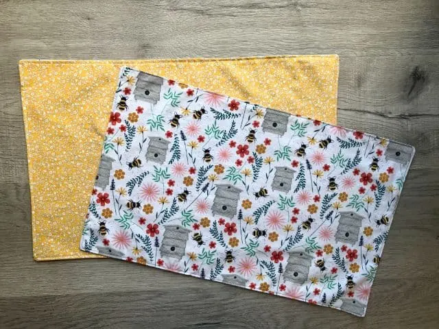 Two fabric placemats in different fabrics - one with mustard yellow flowers and the other with bees, flowers and foliage