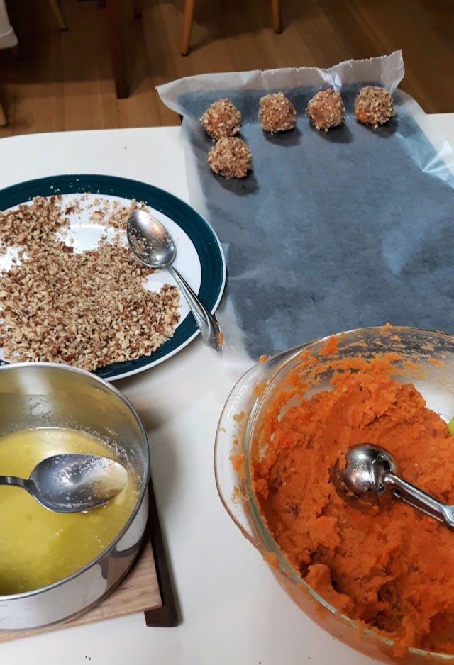 A counter top spread with abowl of mashed sweet potatoes, a pan of melted butter, a plate spread with chopped nuts and a wax paper lined baking sheet with sweet potato balls sitting on top.