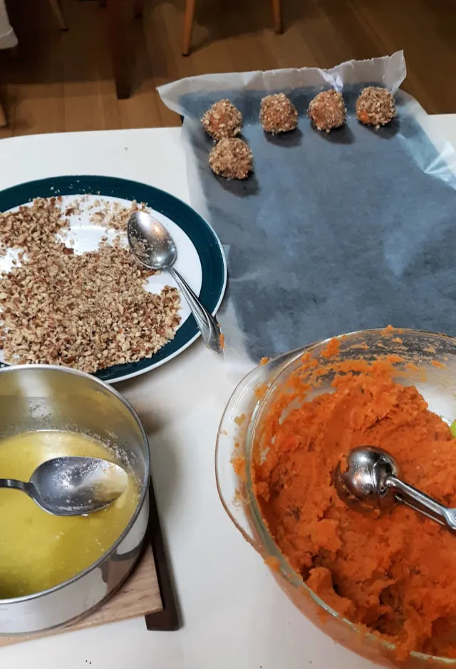 A counter top spread with abowl of mashed sweet potatoes, a pan of melted butter, a plate spread with chopped nuts and a wax paper lined baking sheet with sweet potato balls sitting on top.