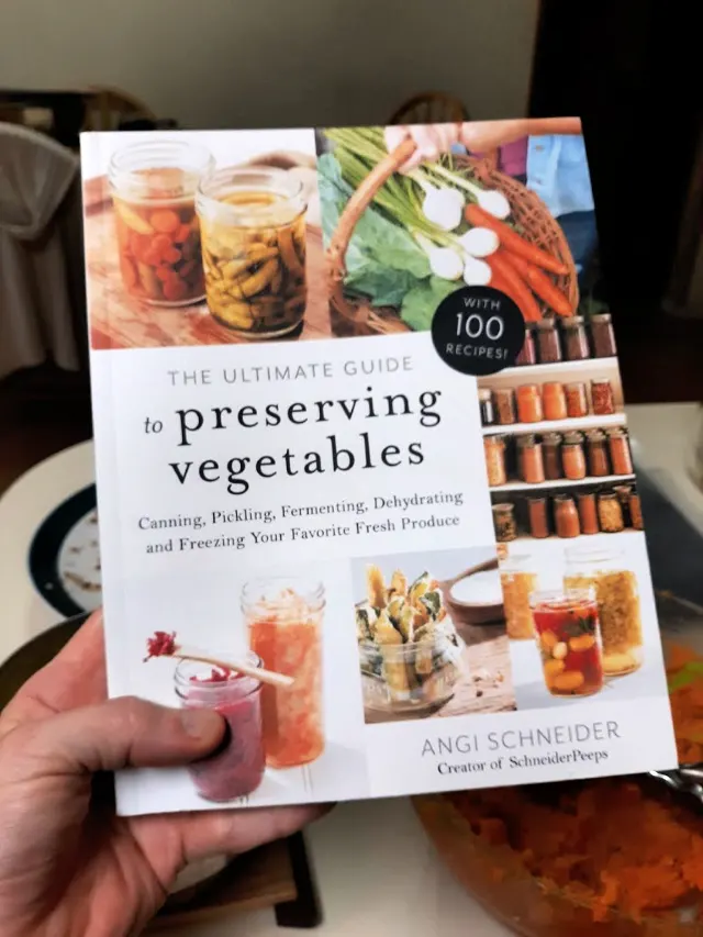 A woman's hand holding a copy of The Ultimate Guide to Preserving Vegetables book.
