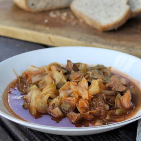 A bowl full of cabbage and beef stew sitting on a table with a cutting board and slices of bread in the background.
