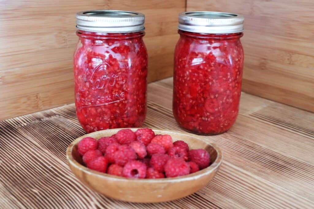 Jars of raspberry pie filling sit on a table behind a wooden bowl full of red raspberries. 