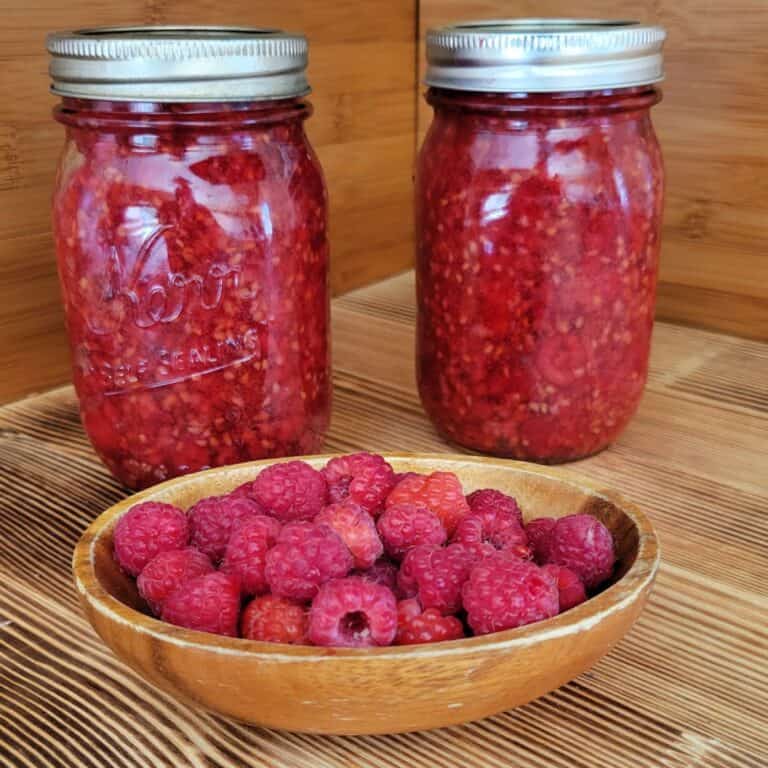2 jars full of pie filling sit on a table behind a wood bowl full of fresh raspberries.