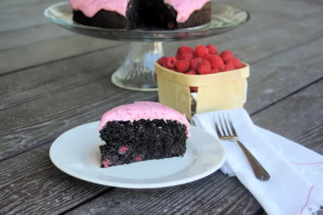A slice of chocolate raspberry cake on a white plate with a napkin and fork sitting to the right - a basket of berries and rest of cake on glass cake plate behind it. 