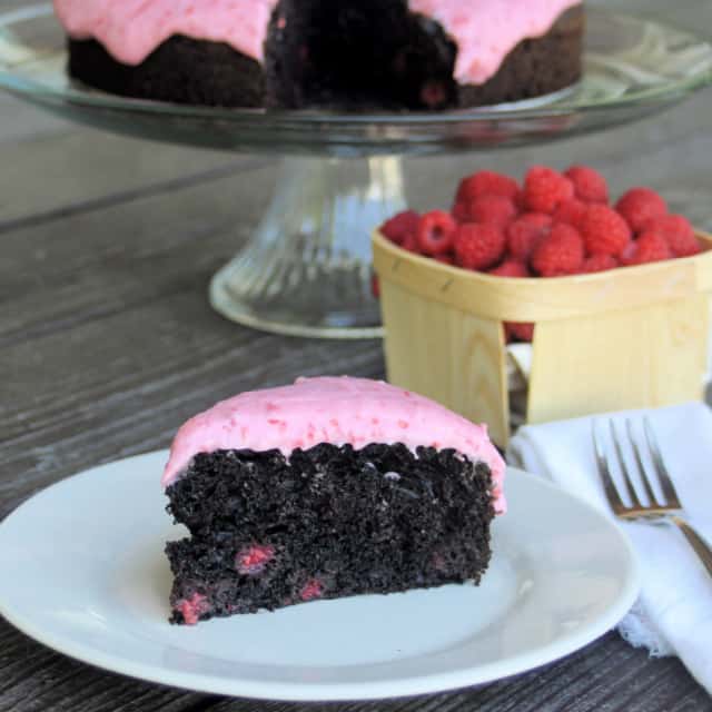 A slice of chocolate raspberry cake on a white plate with a napkin and fork sitting to the right in front of a basket of fresh berries and rest of cake on glass cake plate.