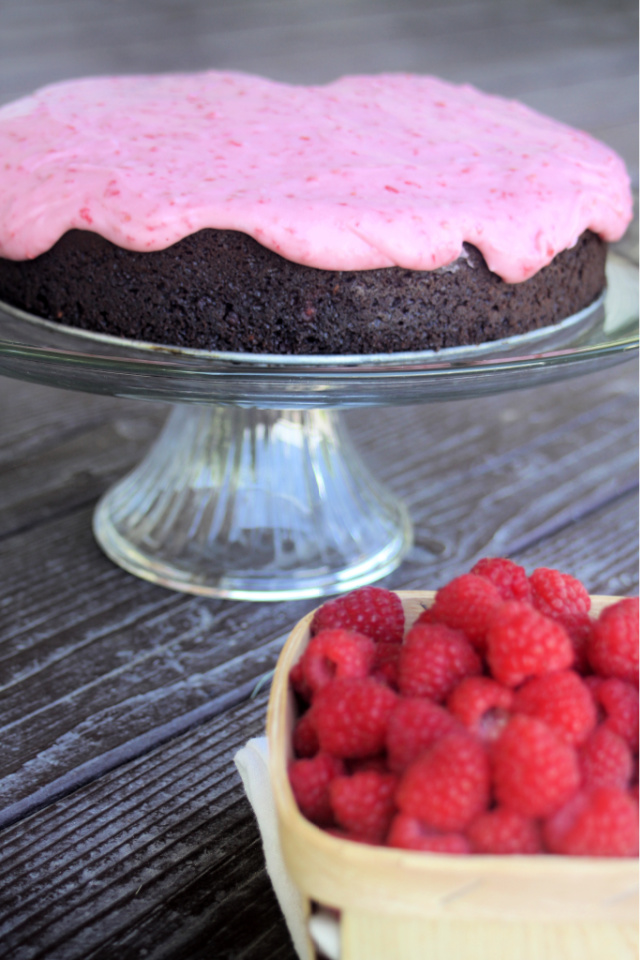 Whole chocolate raspberry cake with raspberry cream cheese frosting on a glass glass plate behind basket of fresh raspberries. 