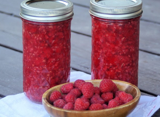 2 jars of raspberry pie filling sitting behind a wooden bowl full of fresh raspberries