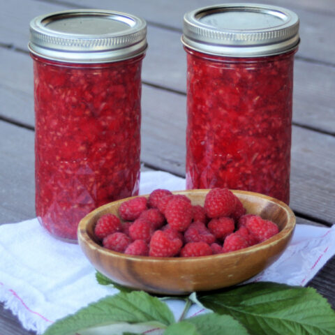 2 jars of raspberry pie filling sitting behind a wooden bowl full of fresh raspberries