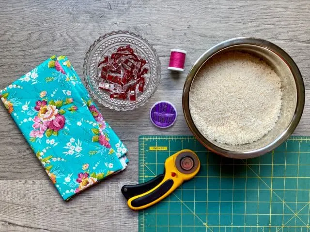 Floral fabric, sewing clips, magenta spool of thread, hand sewing needles, rotary cutter, cutting mat, bowl of rice