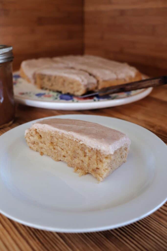 A single frosted apple butter bar sits on a white plate. In the backround more bars sit on a floral cake plate. 