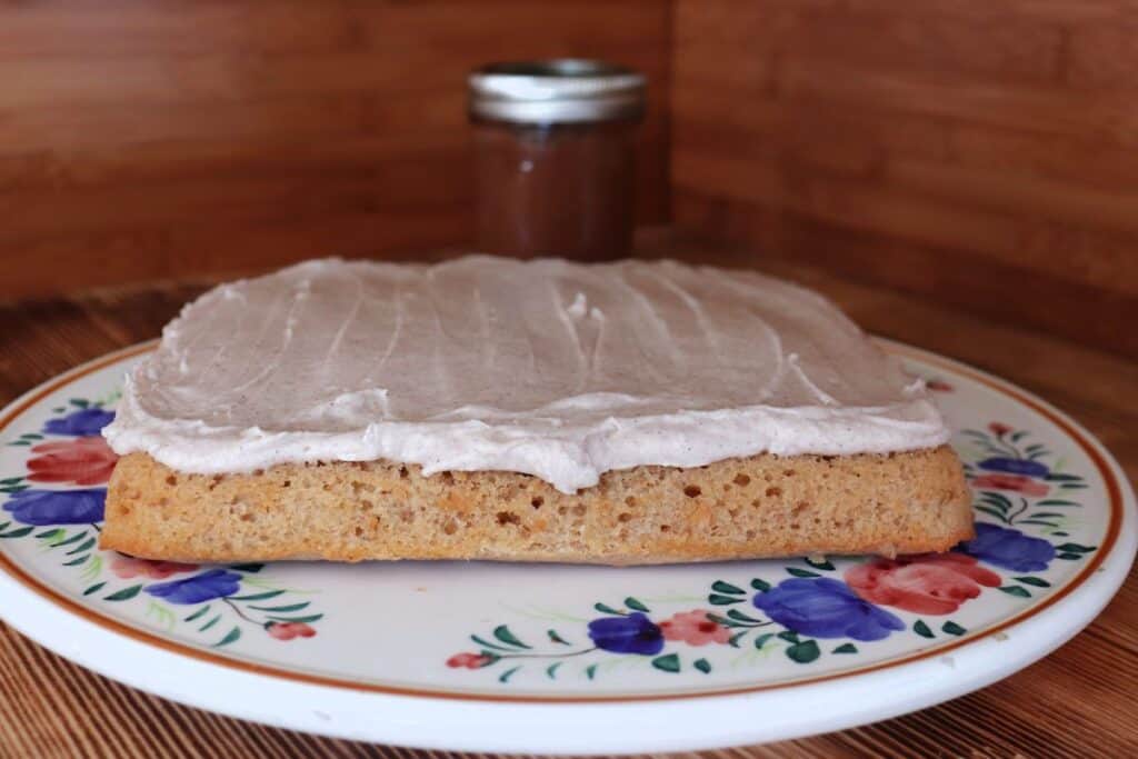Frosted apple butter bars sit, uncut, on a floral cake plate with a jar of apple butter in the background. 