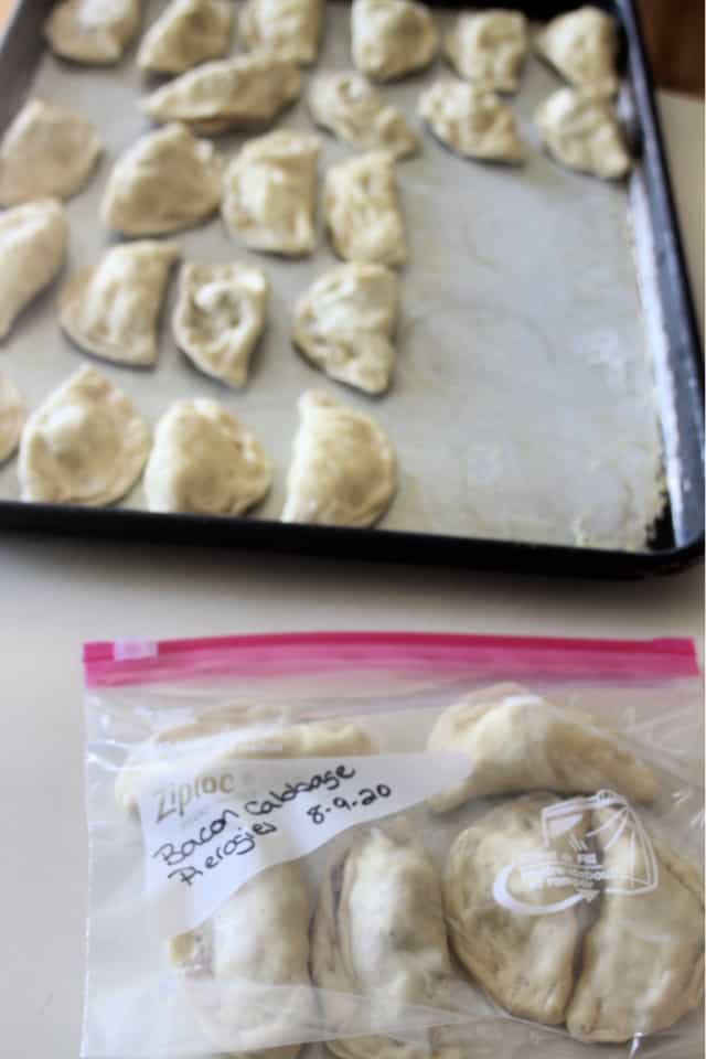 A plastic bag full of frozen bacon cabbage pierogies sitting in front of a cookie sheet full of more frozen pierogies.