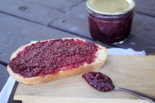 Cherry raspberry jam heaped on a spoon sitting in front of a slice of bread spread with the jam - a sealed jar of jam sitting in the background.