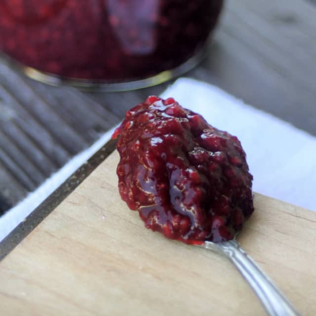A spoon heaped with cherry raspberry jam sitting on a wooden board.