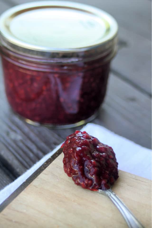 A spoon heaped with cherry raspberry jam on a wooden board with a sealed jar behind it 