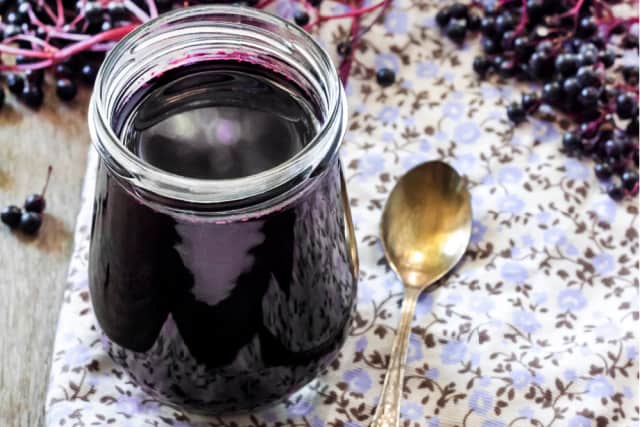 An open jar of elderberry syrup sitting on a floral placemat with a spoon, surrounded by fresh elderberries.