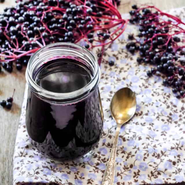 Elderberry syrup in an open glass jar sitting on a floral napkin next to a spoon surrounded by fresh elderberries on the stem.
