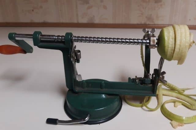 An apple being peeled, cored, and sliced on a green hand crank machine.