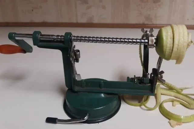 An apple being peeled, cored, and sliced on a green hand crank machine.
