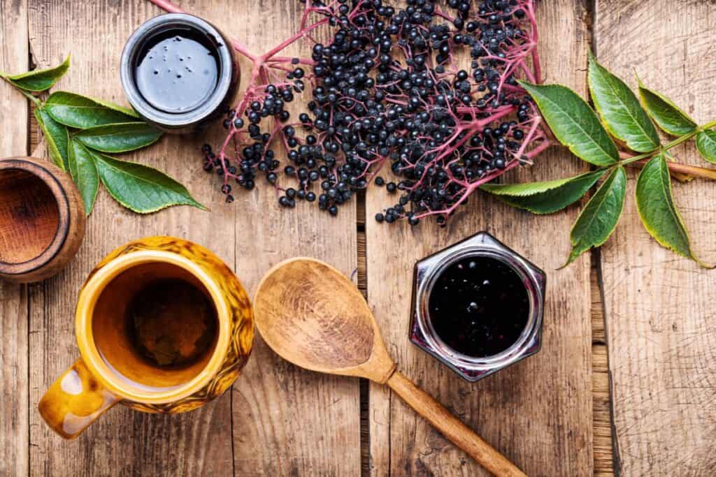 An open jar of elderberry syrup sits on a table surrounded by fresh elderberries, a wooden spoon, and coffee cups. 