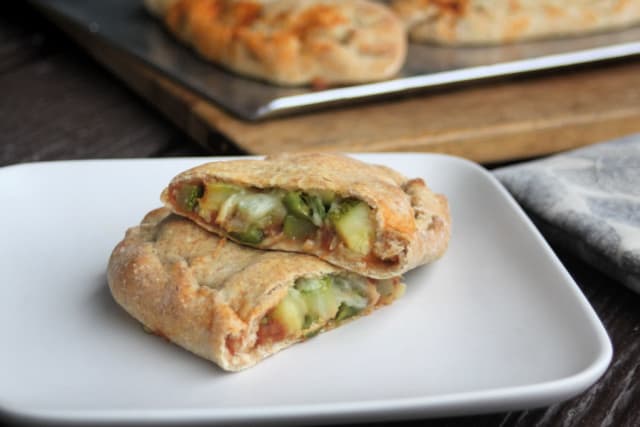 A zucchini calzone cut in half exposing the filling sitting on a white plate with a napkin to the right and a tray of calzones in the background. 