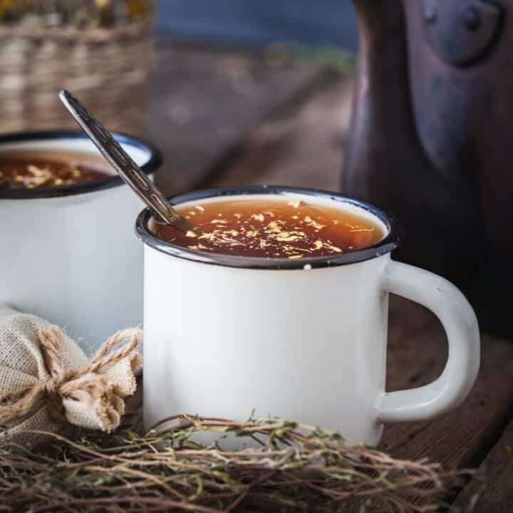 2 white cups full of herbal tea, cup in front has spoon sticking out, surrounded by dried herbs.