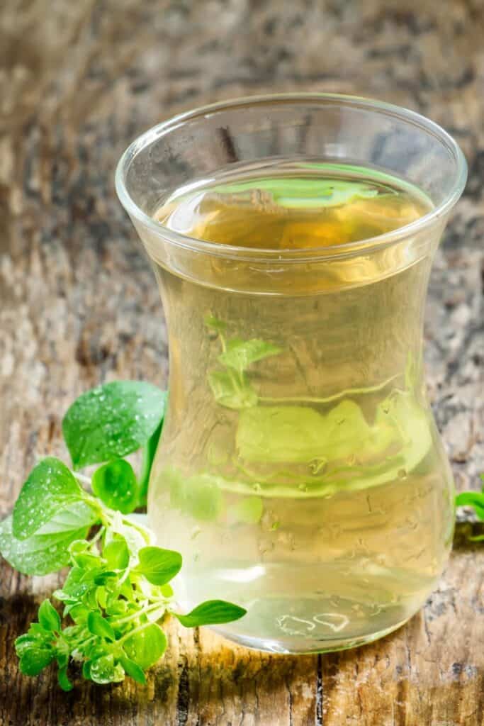 A clear glass full of light colored tea with a sprig of oregano sitting to the left.