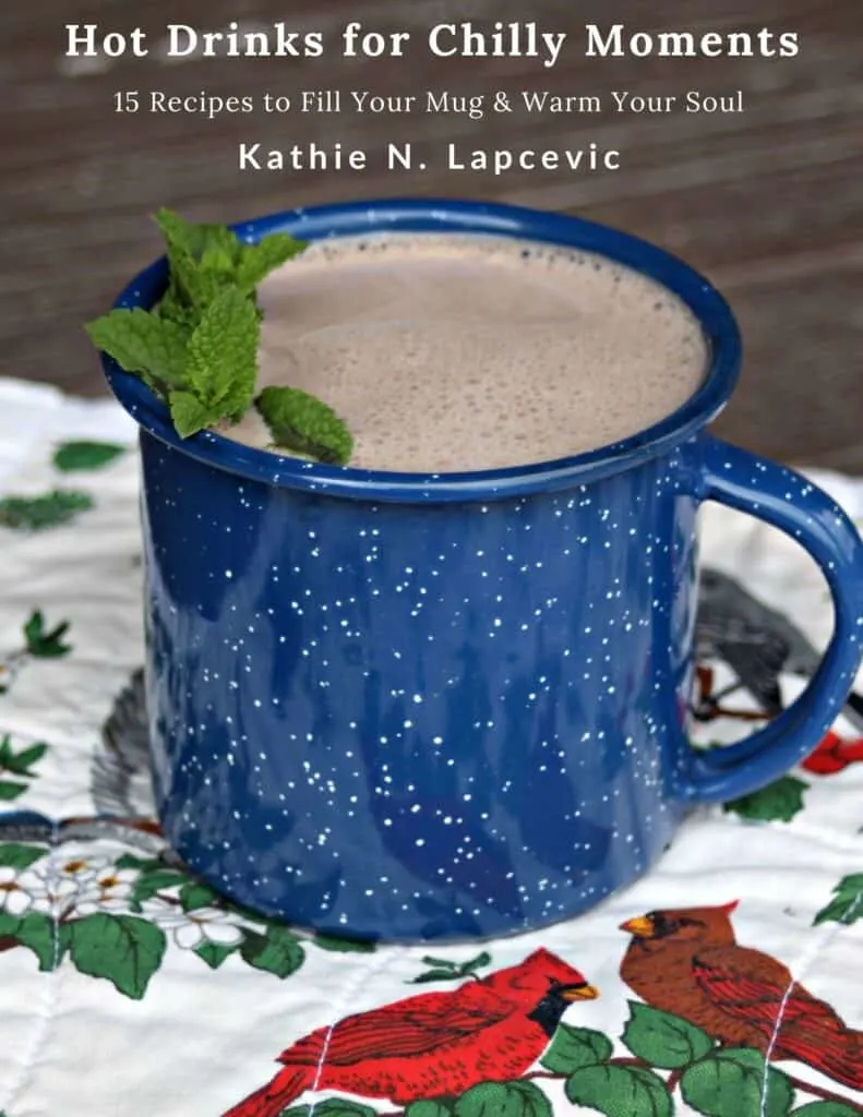 A blue tin cup full of hot cocoa with a sprig of fresh mint sitting on a placemat with cardinals and greenery. Text overlay stating: Hot Drinks for Chilly Moments - 15 recipes to fill your mug and warm your soul by Kathie N. Lapcevic