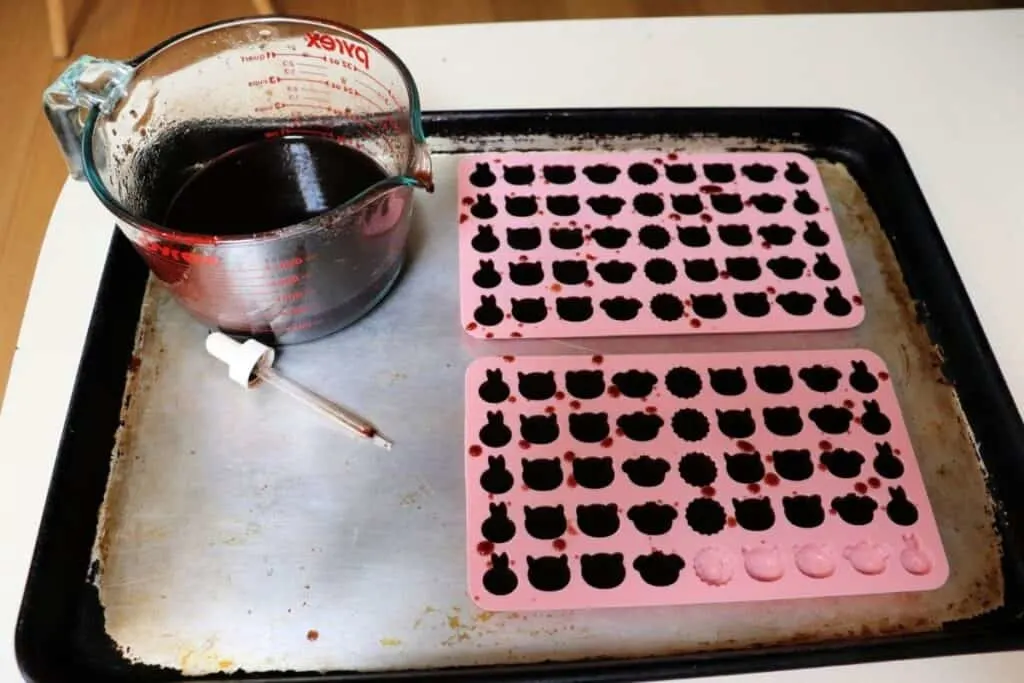 A cookie with 2 pink silicone molds being filled with elderberry syrup gelatin. Sitting next to the molds on the tray is a large glass measuring cup holding more of the liquid gelatin and a small dropper next to it.