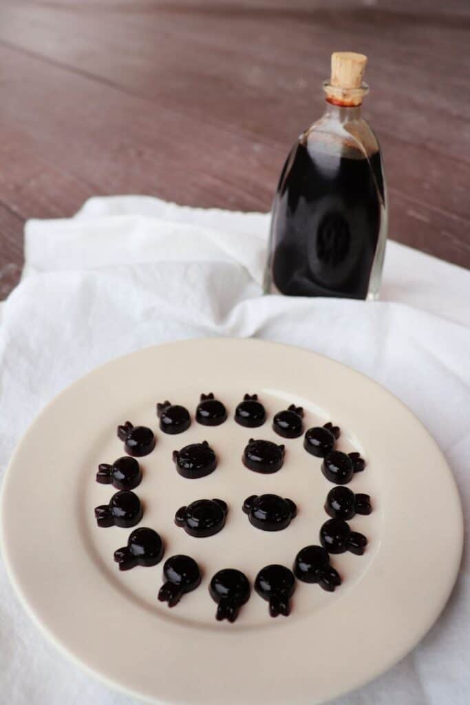 A plate full of elderberry syrup gummies in animal shapes sitting on a white linen with a bottle of elderberry syrup in the background. 