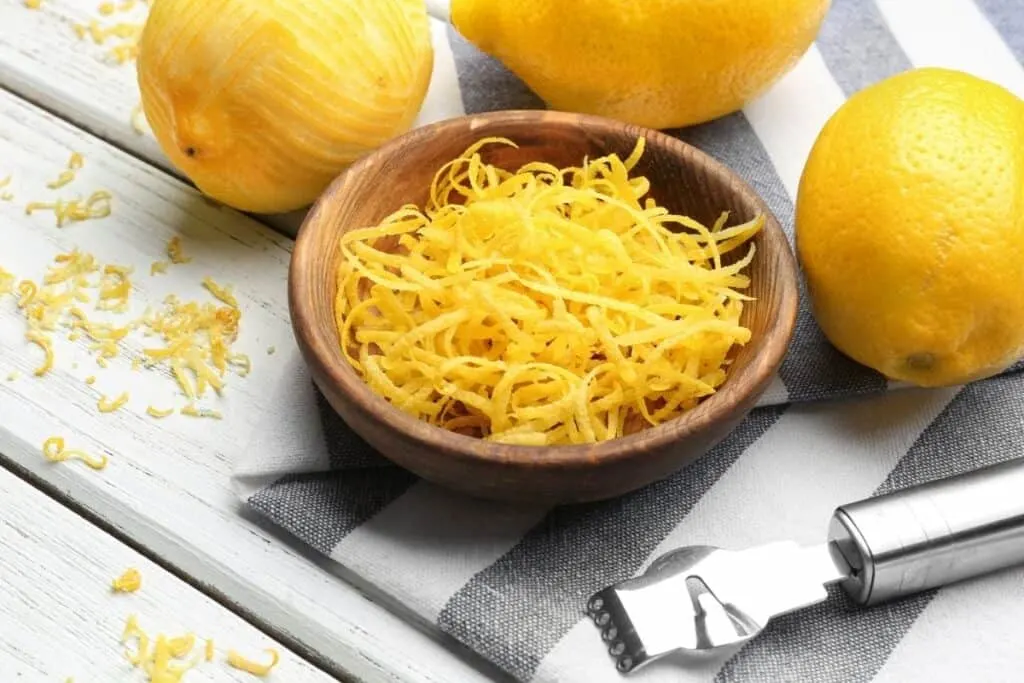 A wooden bowl full of strips of lemon zest surrounded by fresh lemons and steel lemon peeler.