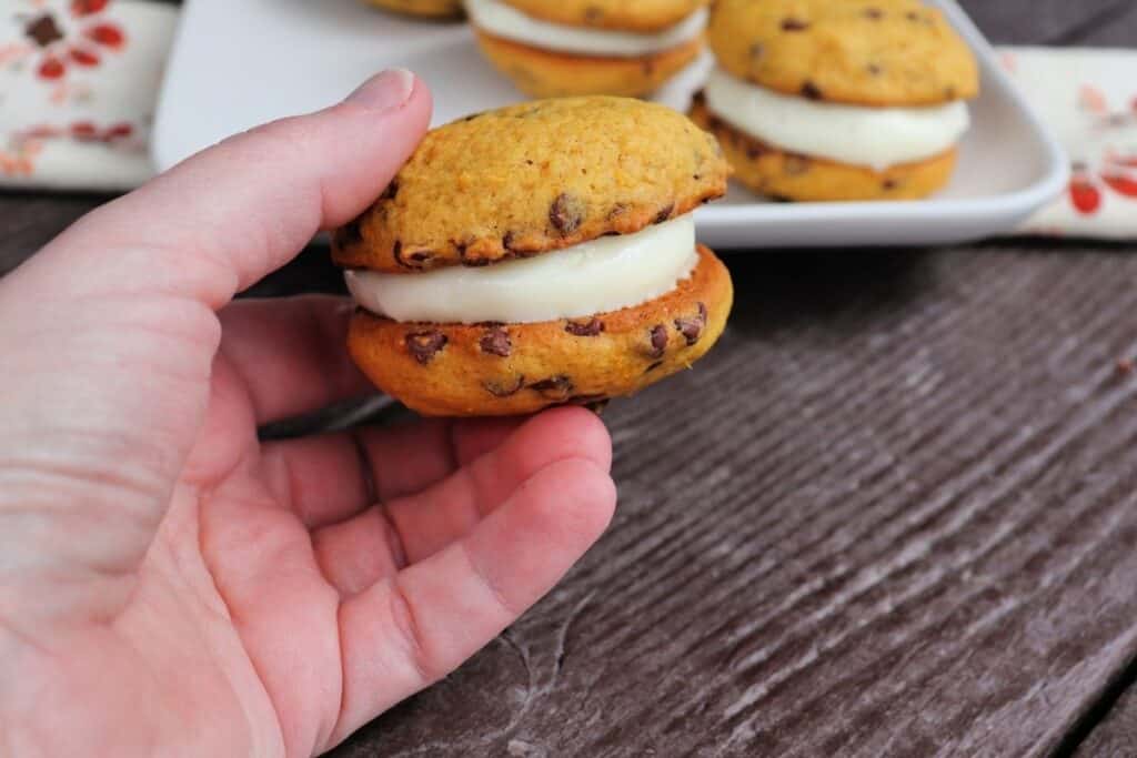 A woman's hand holding a pumpkin whoopie pie with a plate of more whoopie pies in the background.