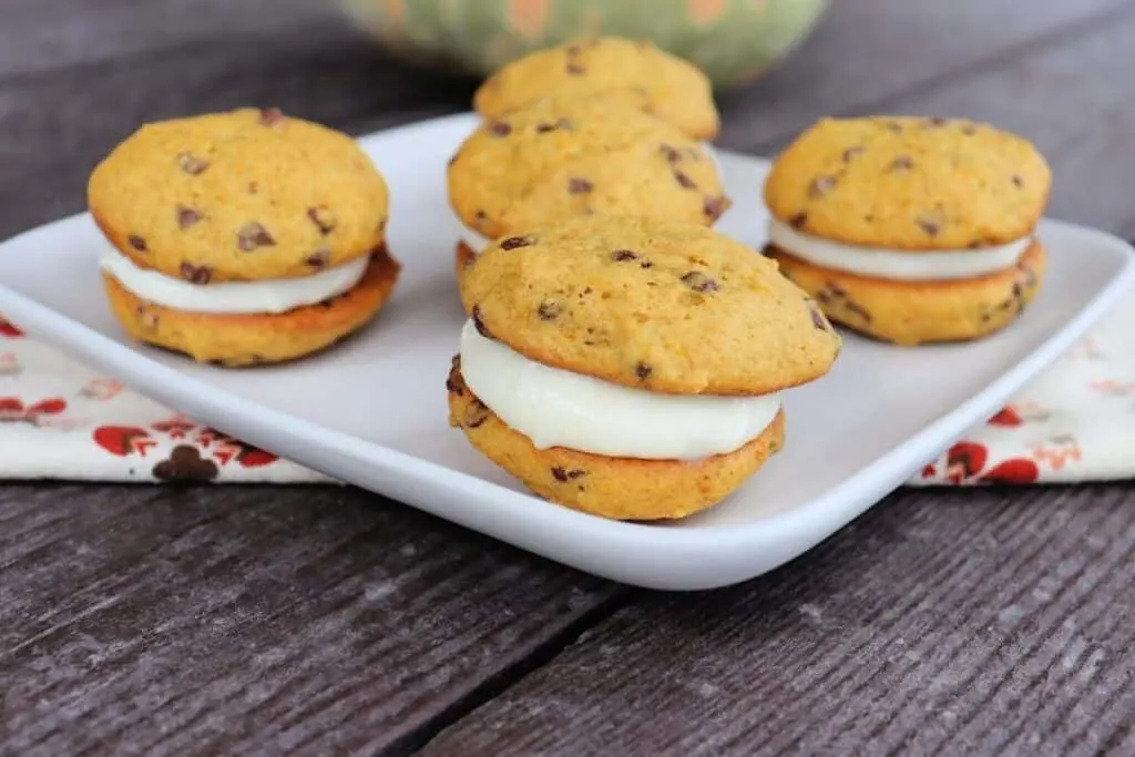A plate full of sandwich cookies with chocolate chips sitting on top of a napkin with a bit of a pumpkin in the background.